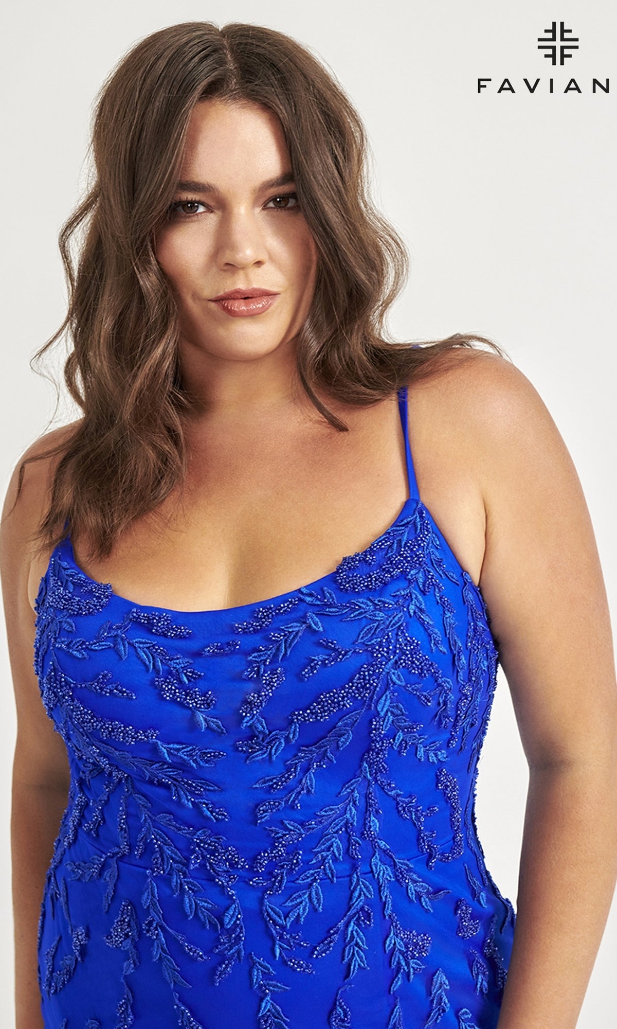 A woman with wavy brown hair wears the Faviana Plus-Size Beaded Long Formal Dress 9559, a sleeveless blue gown with leaf patterns. The